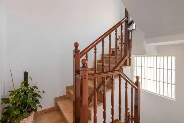 Spacious staircase with marble steps and wooden railings leading to upper floor against backdrop of white walls. Bright daylight from window well illuminates stairs and potted plant on floor