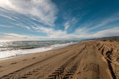Geniş kumlu sahil. Açık mavi gökyüzü ve ince bulutların altında lastik izleri var. Huzuru işaret ediyor..