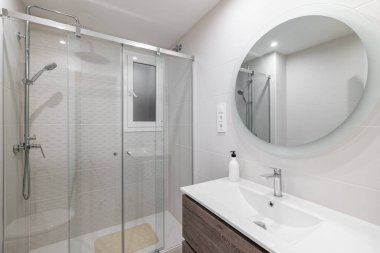 Beyaz seramik fayanslı banyo. Duvarında büyük yuvarlak ayna var. Duş alanını cam parmaklıklar ve metal aksesuarlarla yansıtıyor.