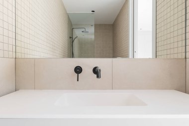 Bej fayanslı modern banyo, beyaz lavabo ve siyah musluk. Aynaya yansıyan cam bölmeli duş alanı.