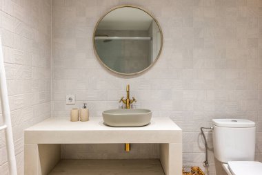 Çok güzel tasarlanmış ve pastel renklerde parlak bir şekilde aydınlatılmış banyoda bakır renkli metal detaylar var. Cam bölmeli duş alanı duvardaki yuvarlak aynaya yansıyor