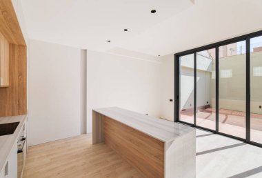 Bol güneş ışığı olan geniş bir mutfak. Modern aletleri olan yemek pişirme alanı, ayrı bir ada şeklinde ahşap masa. Zeminden tavana kadar siyah çerçeveli pencereler çitle çevrili özel terasa bakıyor.