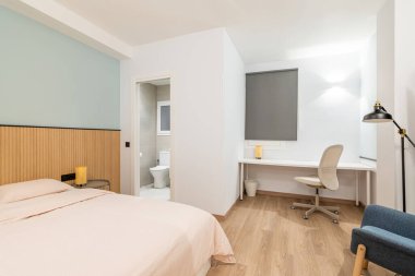 Yatak odası, geniş yatak odası, çalışma alanı, beyaz ahşap masa ve rahat deri ofis koltuğu. Açık kapısı olan küçük özel bir banyo. Yana doğru rahat bir koltuk, yerde lamba var.
