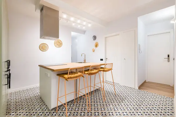 Overlooking large kitchen area. A table in the middle of the room is surrounded by tall bar stools with soft yellow seat cushions. The table has a built-in ceramic hob and a sink with a faucet