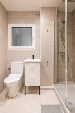 Şeffaf cam duvarla çevrili küçük duş alanı olan banyo. Banyo aksesuarları için çekmeceleri olan mobilyaların üzerinde lavabo. Buzlu cam ve beyaz çerçeveli duvardaki pencere