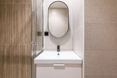 Parlak yapay ışıklandırması olan bir banyo bölmesine yakın çekim, gösteriş için seramik lavabo. Lavabonun üstündeki duvarda büyük modern oval ayna var.