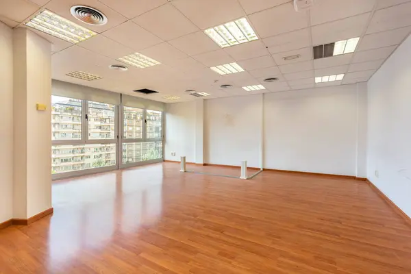 Empty office and fluorescent lamps on the ceiling in need of repair and replacement. Bright office space for rent. Light brown parquet and large panoramic windows in an empty abandoned office