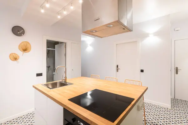 An island table in spacious studio kitchen in modern apartment with trendy authors design. On wooden surface there is an induction electric stove above it with an exhaust system. Mosaic tile floor