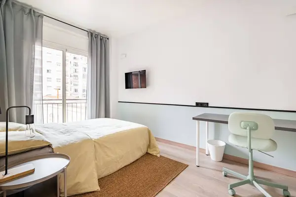 Hotel room with bed for two persons with beige linen. Access to balcony through glass sliding doors. Table against wall with chair for online work. On wall is TV for pleasant evenings watching movie