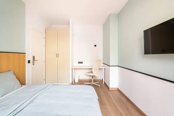 Hotel room with bed for two persons with beige linen. . Table against wall with chair for online work. On wall is TV for pleasant evenings watching movie