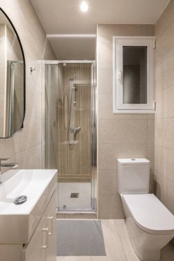 Tavandan parlak ışık saçan küçük bir banyo. Duvarlarda bej granit fayanslar. Beyaz klozet ve lavabo tuvalet masasının üzerinde, şeffaf cam bölmeli duş alanı. Oval ayna duvarda