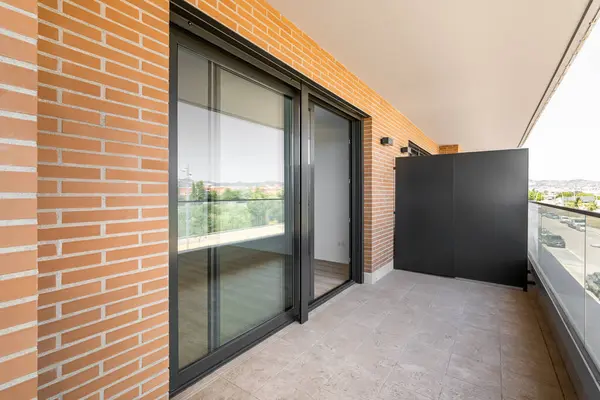 Large entrance balcony door made of black plastic with a sliding system. Terrace with marble floor and glass railings. The walls of the building are made of yellow bricks