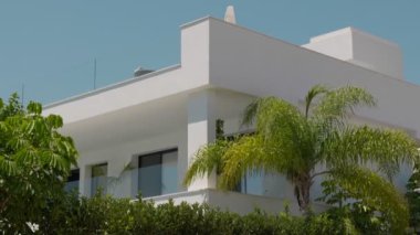 Beyaz minimalist bir villa, yemyeşil palmiyelerden oluşan bir arka fonda duruyor, yapraklar rüzgarda hafifçe çırpınıyor..