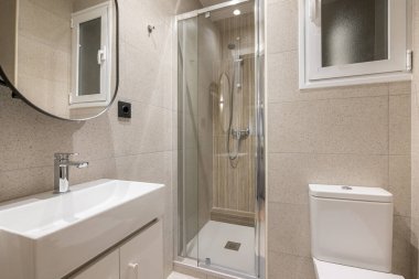 Cam duş kabini olan bej fayanslı şık kompakt banyo, beyaz tuvaletli lavabo ve ayna. Otelde ya da dairede küçük ama çok işlevli banyo kavramı.