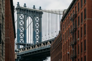 New York 'taki iki tarihi kırmızı tuğla bina arasındaki Manhattan Köprüsü' nün pitoresk bir görüntüsü. Sahne, şehir mimarisini ve köprünün ihtişamını yansıtıyor..