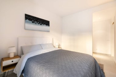 Çift kişilik yataklı, duvarında resim olan bir yatak odası ve açık renkli komodinlerle süslü komodinleri ve koridora açılan açık bir kapı. Kompakt otel odası kavramı. Kopyalama alanı.