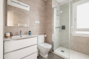 Tuvaletli cam duş kapılı bej fayanslı banyo ve ayna ve gün ışığı pencereli lavabo. Modern banyo tasarımı konsepti.