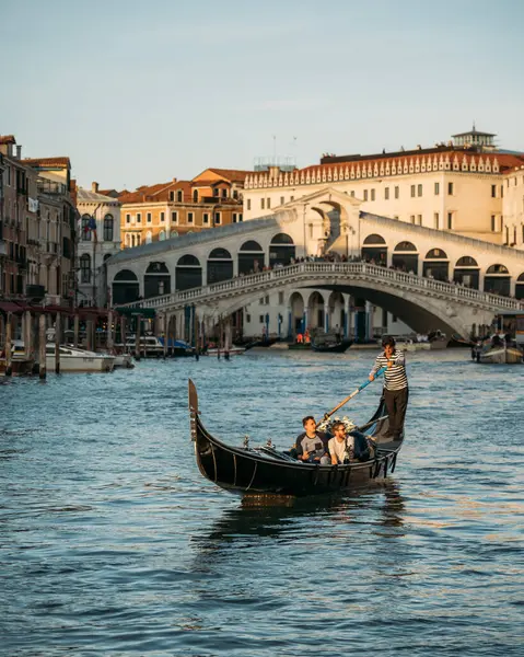 30 Eylül 2021, VENECIA, İtalya: Güneşli yaz akşamında Rialto Köprüsü 'nün arka planında gondolcuyla gondolda oturan turistlerin güzel manzarası