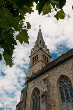 Lihtenştayn 'daki yeşil yapraklarla çerçevelenmiş parçalı bulutlu gökyüzüne karşı yükselen güzel bir kilise kulesi..