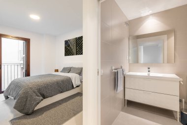 Samimi bir banyodan, şık bir bej tonda, içinde lavabo ve parlak bir odada yatağı olan yatak odasına bakan bir ayna olan bir görüntü. Şık minimalist tasarım konsepti.