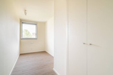 Sağ tarafında gardırop olan boş, küçük, parlak bir yatak odası. Mobilyasız pencereli minyatür çocuk odası