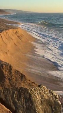 Gün batımında yumuşak dalgaların kayalık kıyılara hafifçe vurduğu sakin ve huzurlu bir sahil manzarası.