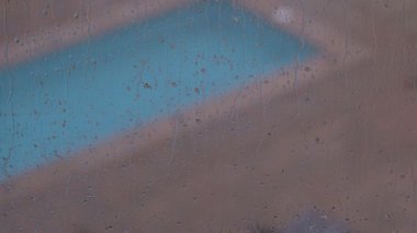 Pencereye doğru akan yağmurun yakın çekimi, mavi renkli bir yüzme havuzunun görüntüsünü bozuyor..