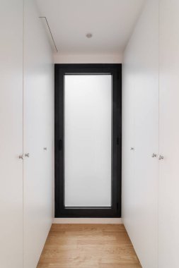 Temiz beyaz dolapları olan parlak ve minimalist bir koridor tasarımı, siyah çerçeveli buzlu bir cam kapı ve doğal ahşap döşeme..