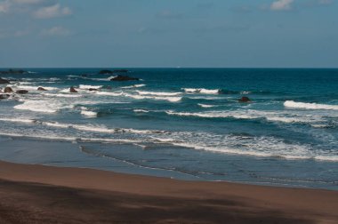 Köpüklü dalgaların olduğu manzaralı bir deniz manzarası kayalık bir plajda yuvarlanıyor, bulutlar ve mavi gökyüzü karışımı.