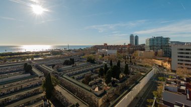 Barselona yeşil park alanlarındaki tarihi mezarlığı ve güneşli gökyüzünün altındaki kıyıya yakın modern gökyüzü binalarını birleştiren geniş bir manzara.
