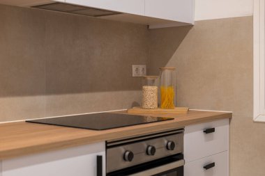 Ahşap tezgah, beyaz dolaplar, elektrikli soba ve tahıl ve makarna dolu kavanozları gösteren modern bir mutfak.