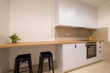 Beyaz dolapları, uzun ahşap tezgahı, siyah tabureleri ve paslanmaz çelik fırını olan modern bir mutfak.