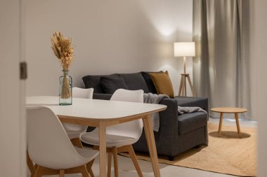 Beyaz sandalyeler, kurumuş çiçekler ve gri koltuklu mavi bir vazo içeren parlak bir yemek ve yaşam alanı.