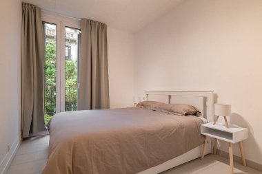 Minimalist yatak odası, çift kişilik yatak örtüsü, beyaz mobilya ve perdeli büyük bir pencere.