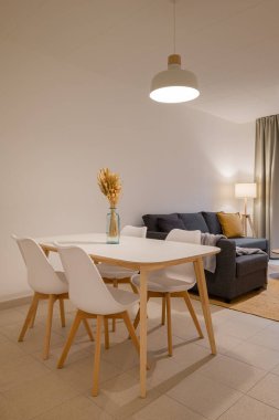 Parlak bir odada beyaz bir masa, tahta bacaklar, dört sandalye ve vazo var.