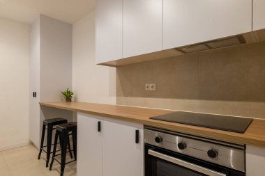 Mutfak perspektifi ahşap tezgah, beyaz dolaplar, fırın, siyah tabure ve minimalist tarzda küçük bitkileri gösteriyor.