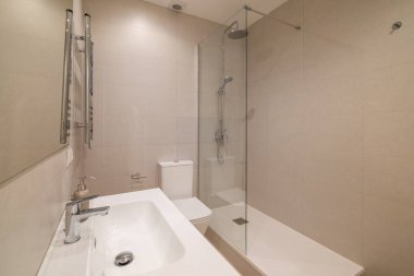 Modern banyo içi bej kaplamalı duvarlar ve cam duş perdesi ve krom armatürlerle döşenmiş zemin.