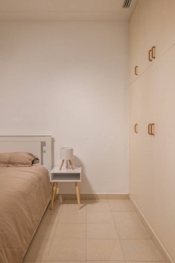 Küçük yatak odasının köşesinde boz yatak örtüsü, komodin ve doğal tonlarda gardırop var.