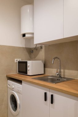 Mutfak köşesinde pencere, ahşap tezgah, beyaz dolaplar, mikrodalga fırın, çamaşır makinesi ve lavabo var.