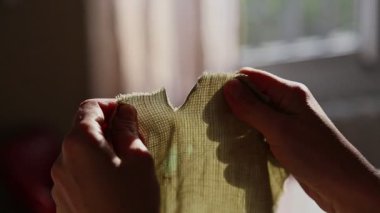Toz parçacıkları güneş ışığıyla aydınlanırken, yıpranmış kenarları ortaya çıkaran, dokulu bir kumaşı ayıran bir çift el..