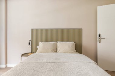 Yatak odasının içinde beyaz bej yastıklı çift kişilik bir yatak, kaburgalı bir yatak başlığı yuvarlak bir masa ve bir lamba var..