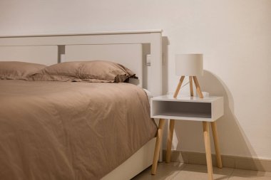 Işıl ışıl ve düzenli bir yatak odası, bembeyaz yatak çerçevesi ve lambalı basit bir ahşap komodin.