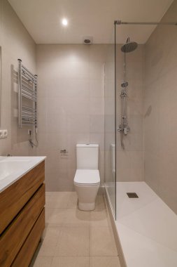 Modern bir banyo krom havlu ısıtıcısı, ahşap süs ünitesi ve bej kiremitlerle kaplı cam duş muhafazasını gösteriyor.
