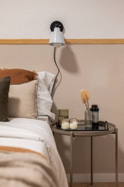 Yatak odasının köşesinde bej yatak ve yastıklar var. Yanında da küçük yuvarlak bir masa, mumlar ve dekoratif vazolar var..