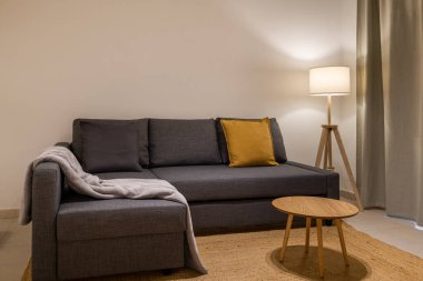 Gri L şeklinde kanepesi, sarı yastığı, ahşap masası ve yumuşak bir lambası olan basit ve zarif bir oturma odası.