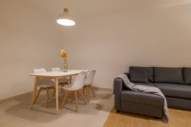 Beyaz bir masa ve minimalist bir ortamda rahat gri bir kanepeye bitişik sandalyeli kompakt yemek köşesi.