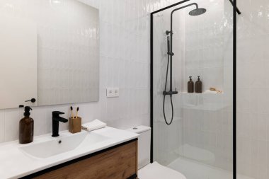 Cam duşlu modern banyo, beyaz fayanslar, ahşap kibir ve siyah eşyalar. Tezgahında tuvalet malzemeleri olan minimalist tasarım..