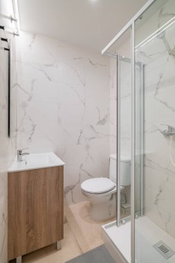 Cam banyosu, beyaz tuvaleti ve ahşap kibiri olan modern bir banyo. Mermer desenli duvarlar parlak, çağdaş bir görünüm yaratır.