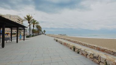 Palmiye ağaçları ve bulutlu bir gökyüzünün altında kumlu bir plajla kaplı bir sahil geçidi. Sahne sakin ve davetkar, sakin yürüyüşler için mükemmel..