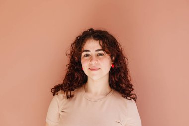 Kıvırcık saçlı ve çilli genç bir kadın kameraya doğal bir ifadeyle bakıyor. Turuncu pembe arkaplan